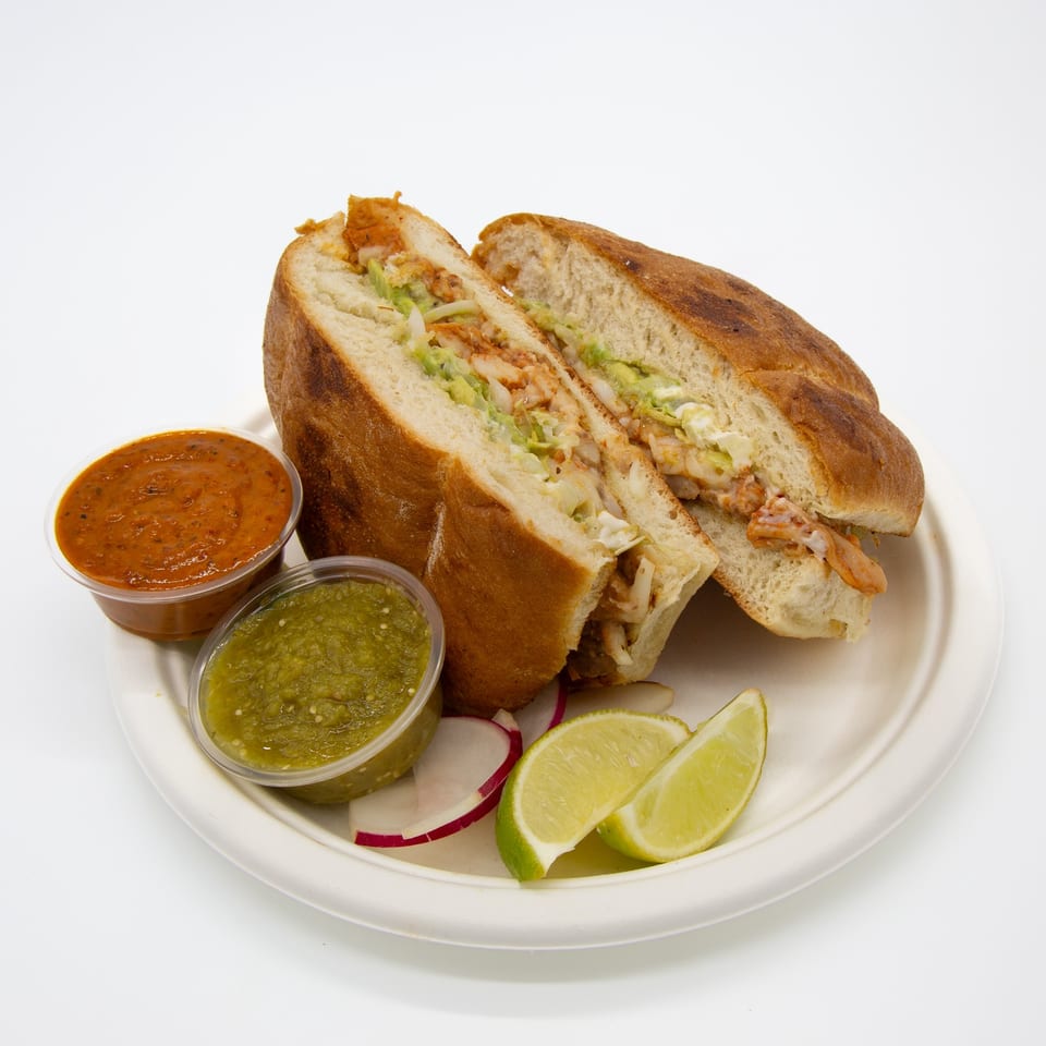 A Mexican torta sandwich, cut in half, with two salsa cups, lime wedges, and radish slices.