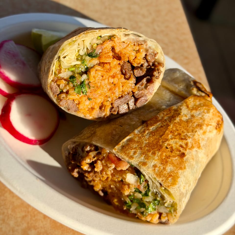 Two Studio Tacos burritos, one cut in half, revealing rice, meat, and vegetables, with radishes and lime.