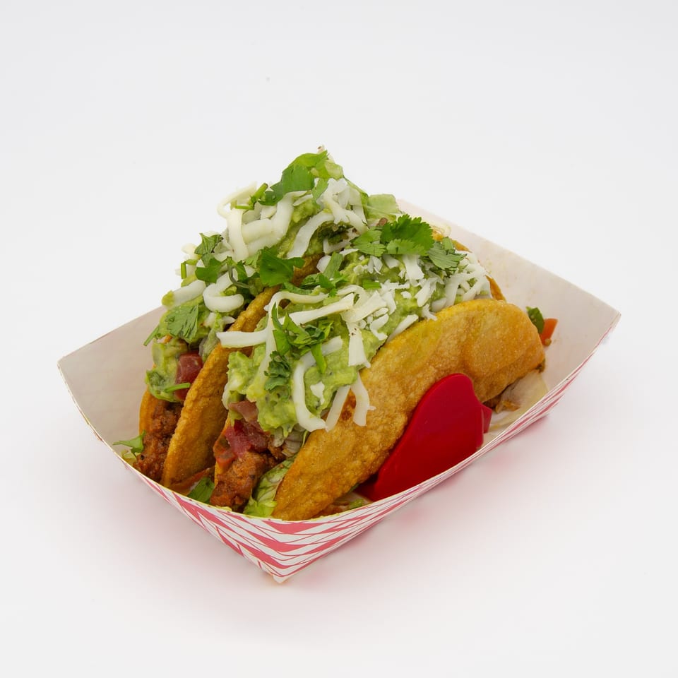 Three Studio Tacos with guacamole, shredded cheese, and cilantro in a paper tray.